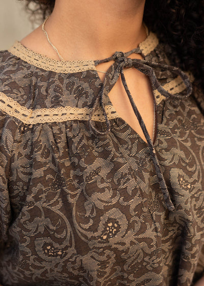 Close up of the fabric and gold trim on the brown pattern top with a tie at the neck.